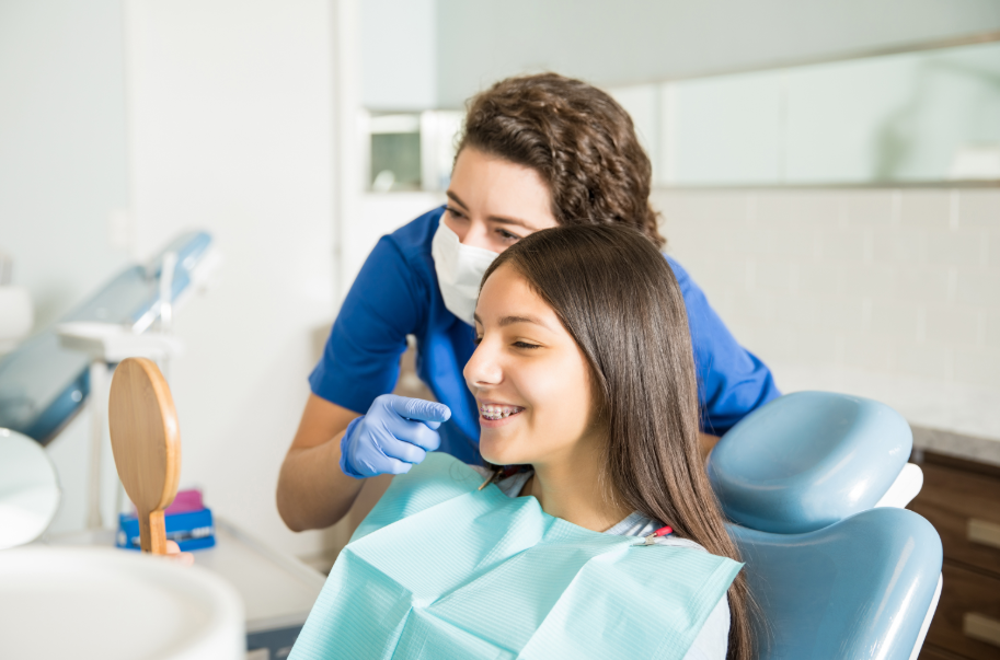 Teen girl with braces in dental chair admiring smile in mirror with dentist pointing to teeth